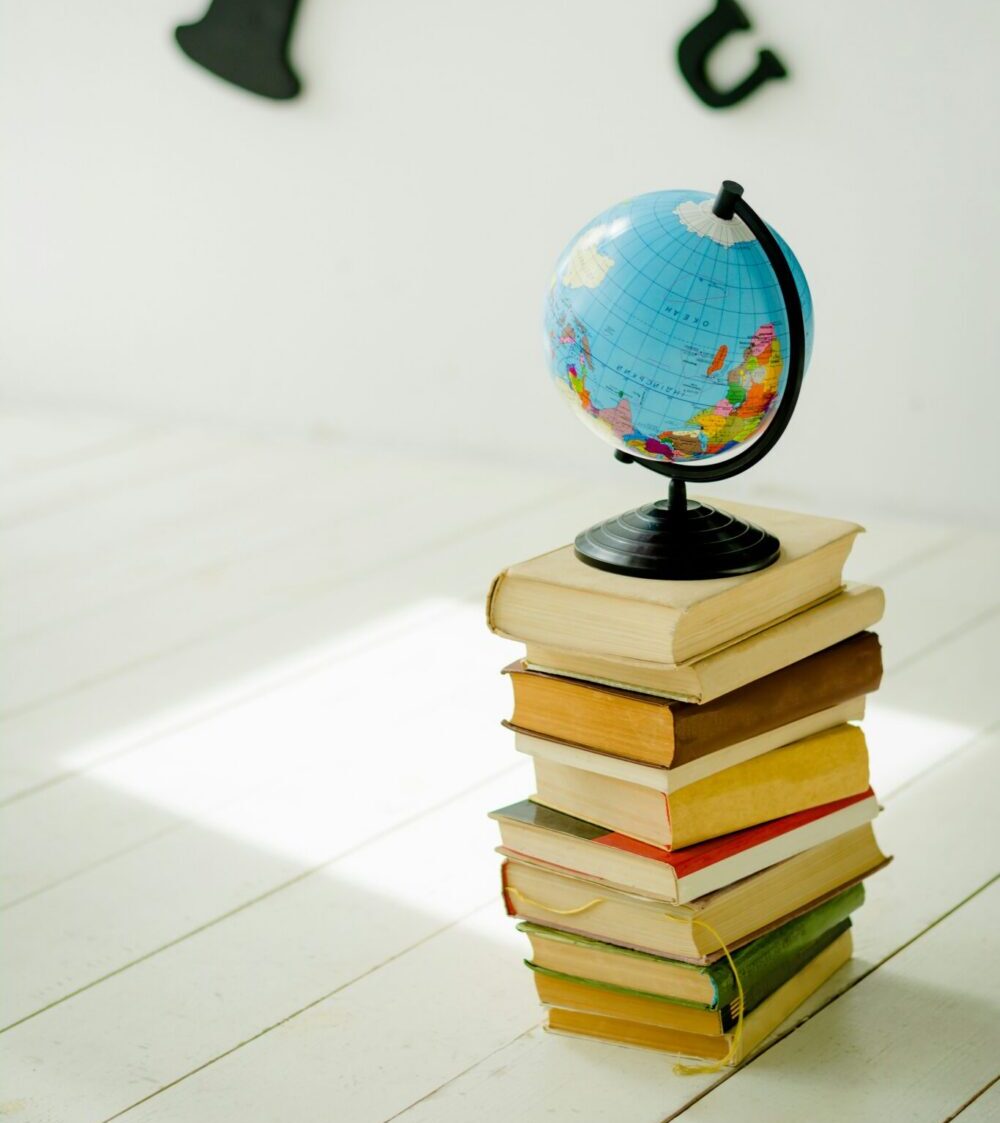 A small globe resting on top of a tall stack of colorful hardcover books placed on a white wooden floor, with large black letters blurred in the background.