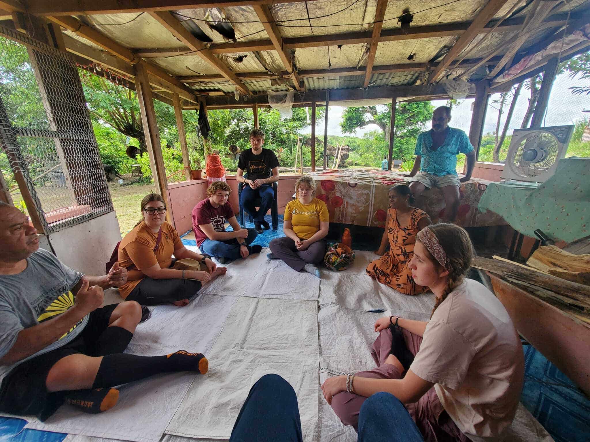 Group of people seated in a circle on floor mats inside an open-air wooden structure with a corrugated roof, surrounded by greenery, while one person stands near a table with a fan and household items.
