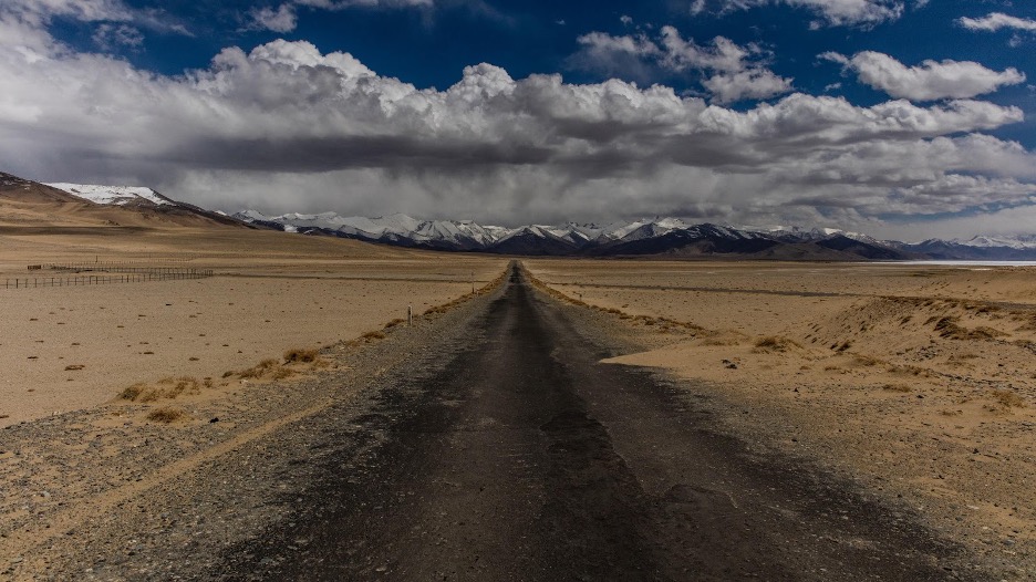 A long, straight dirt road stretches through a vast, barren landscape toward distant snow-capped mountains.