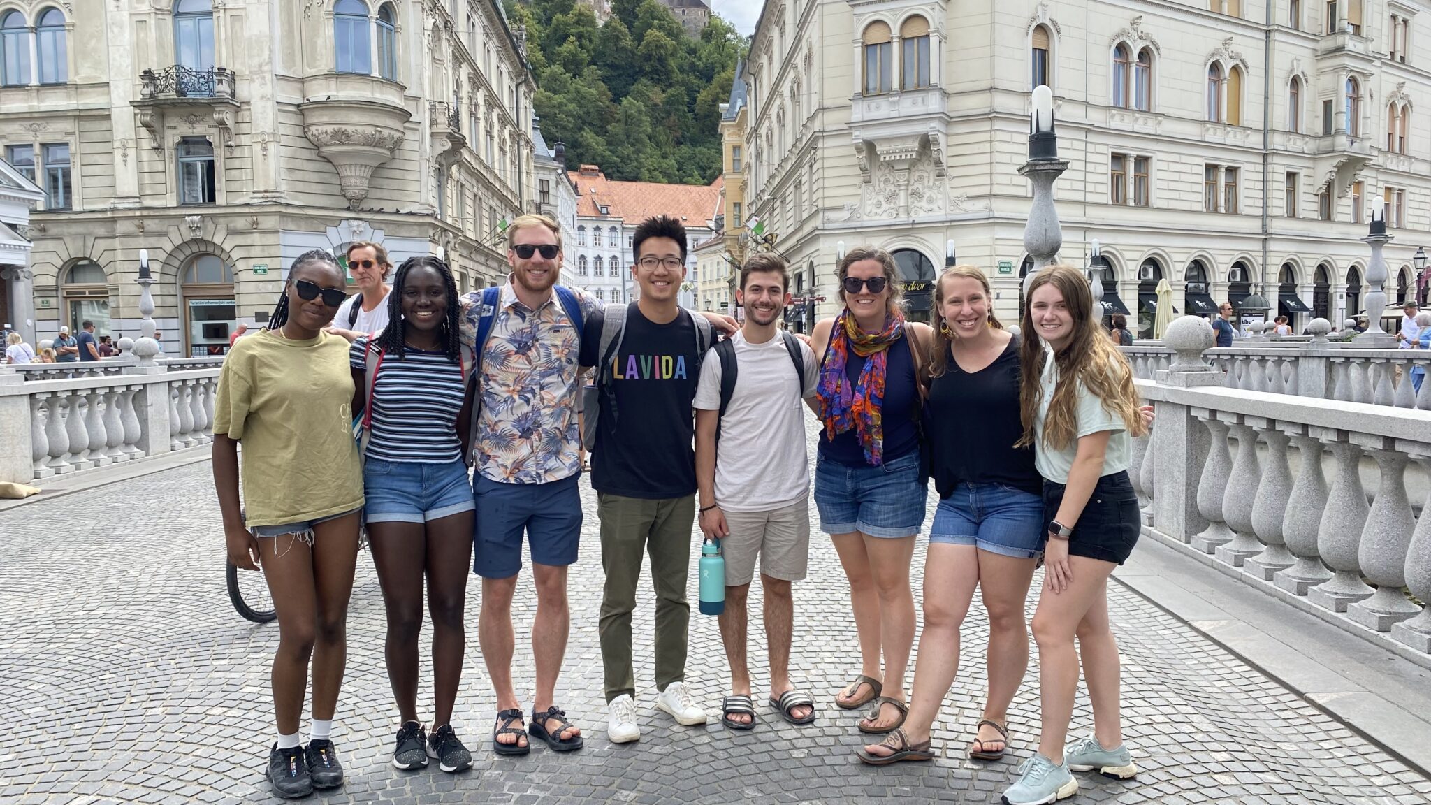 Group of eight friends standing close together on a cobblestone bridge in a European city, smiling at the camera.