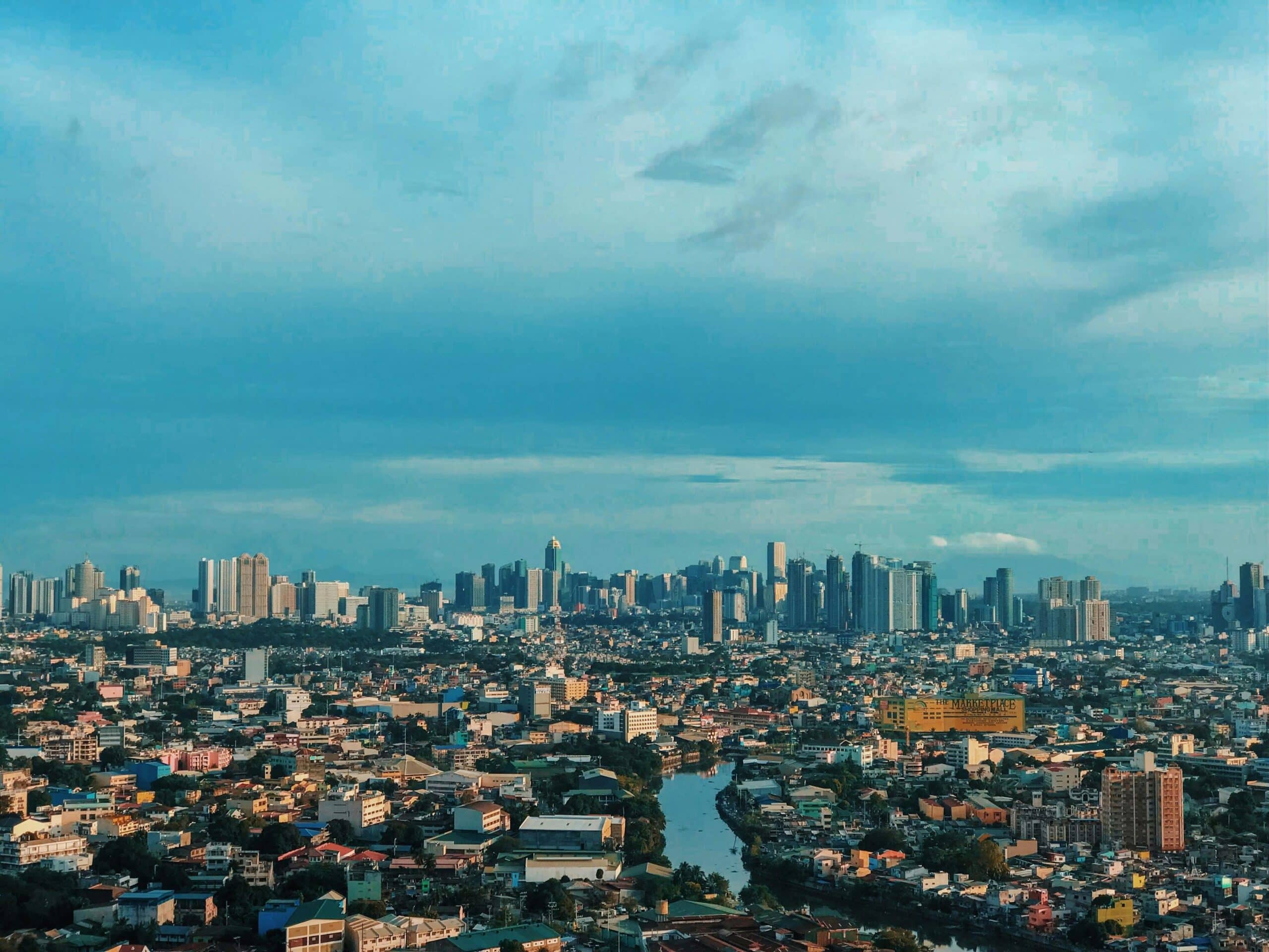 Panoramic city skyline along a river under a clear blue sky, with numerous buildings stretching across the horizon.