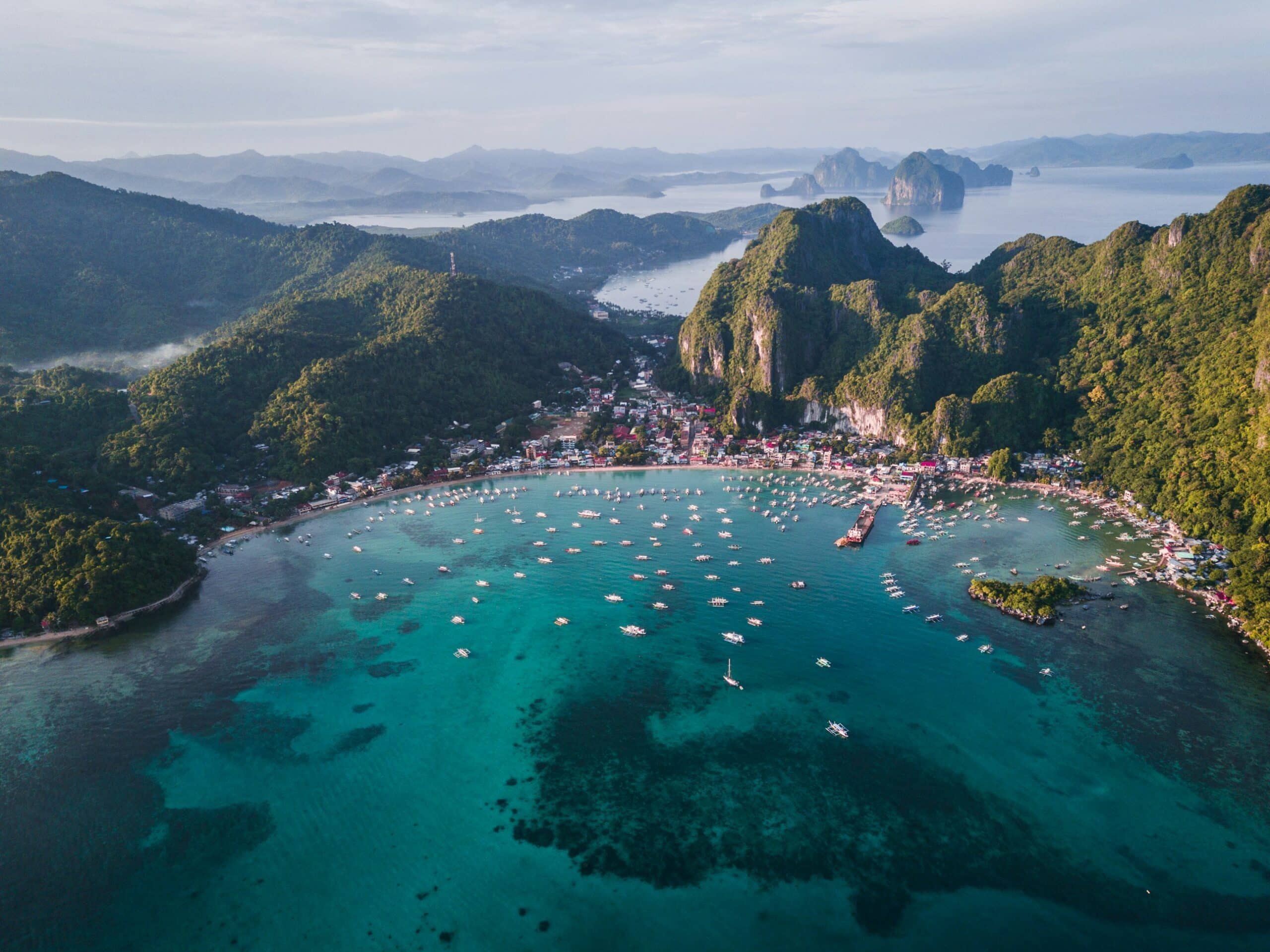 Aerial view of a turquoise bay with a small town along the shore and hundreds of boats, set among lush green hills and distant islands.