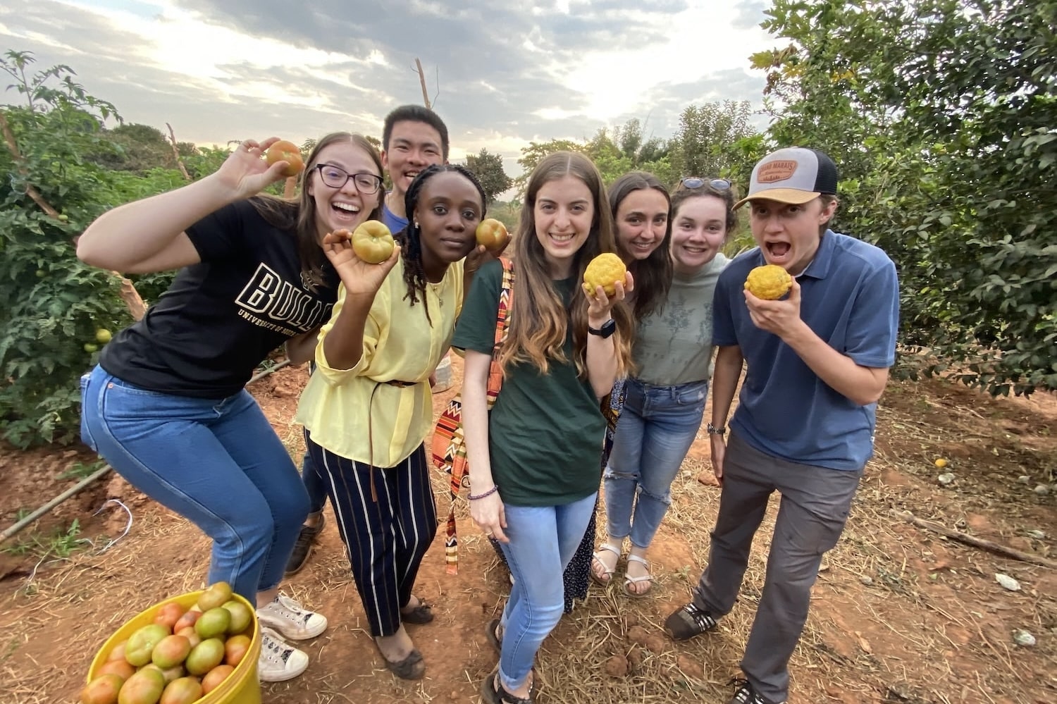 Seven smiling friends in an orchard pose with freshly picked apples, one holding a yellow fruit, bucket of fruit on the ground nearby.