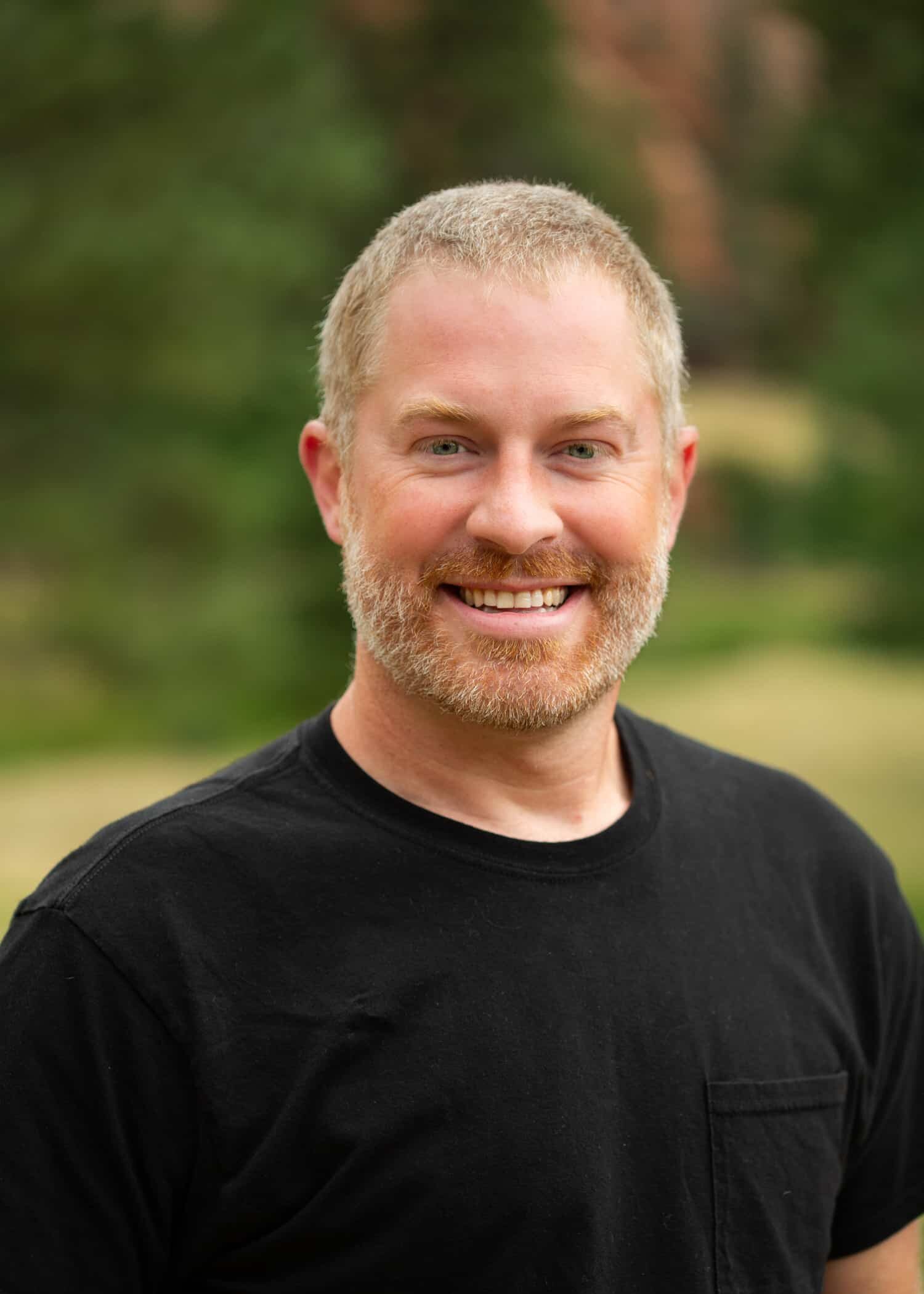 Close-up portrait of a smiling man with short blond hair and a light beard, wearing a black T-shirt, outdoors.