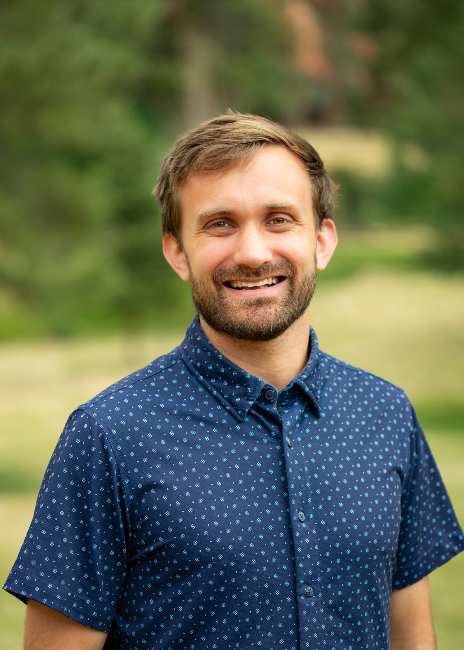 Smiling man outdoors wearing a navy blue shirt with small white dots, natural green background behind him.