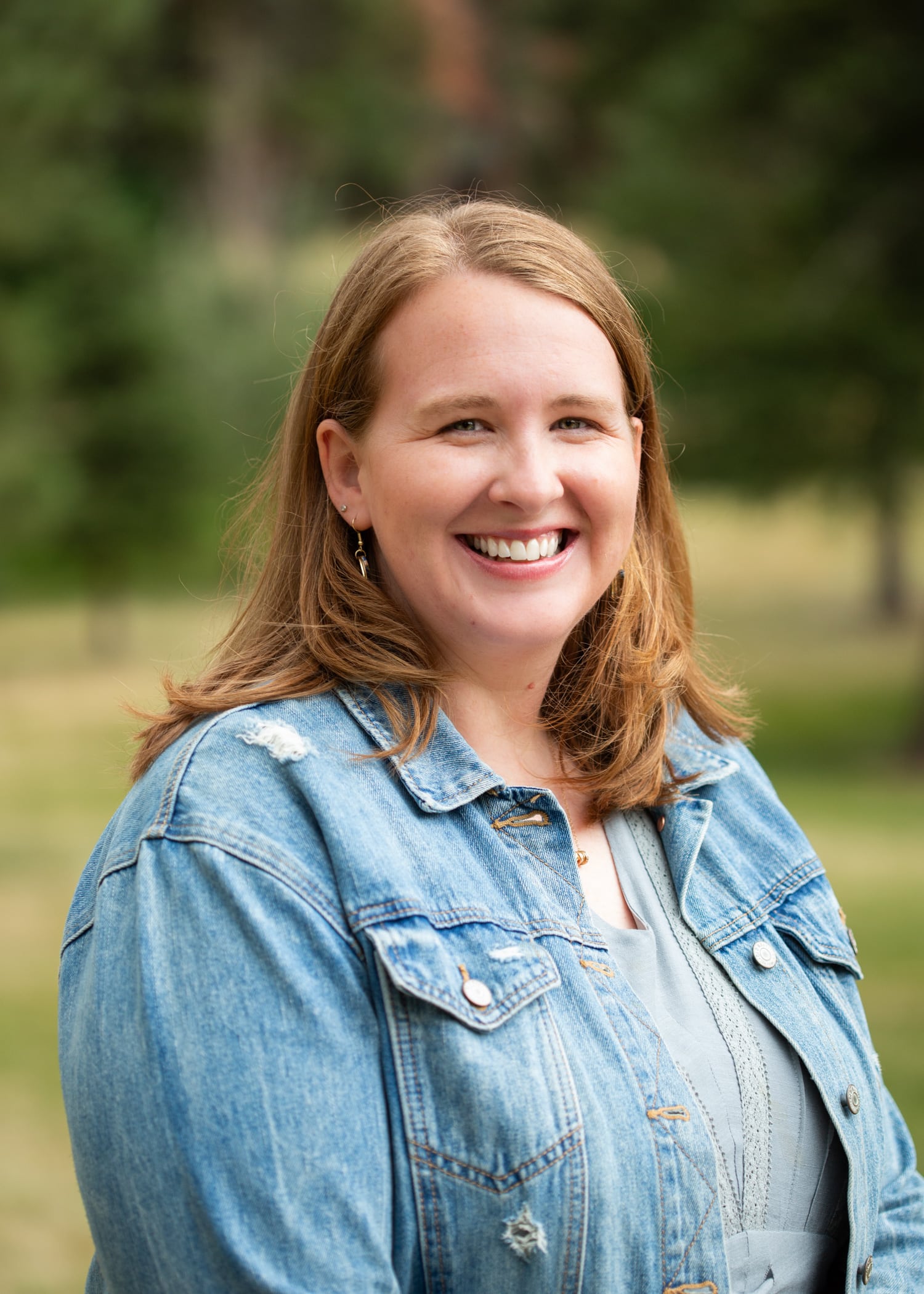 Smiling woman outdoors with light brown hair, wearing a denim jacket and gray top.