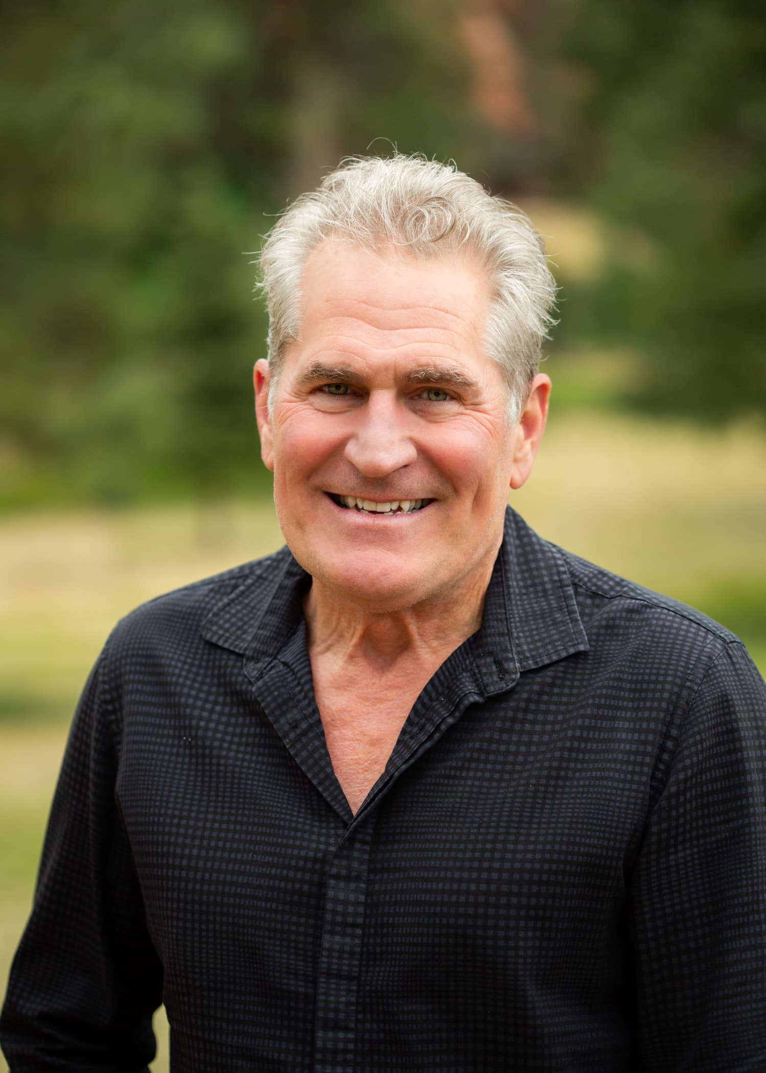 Smiling middle-aged man with gray hair outdoors, wearing a dark checkered shirt.