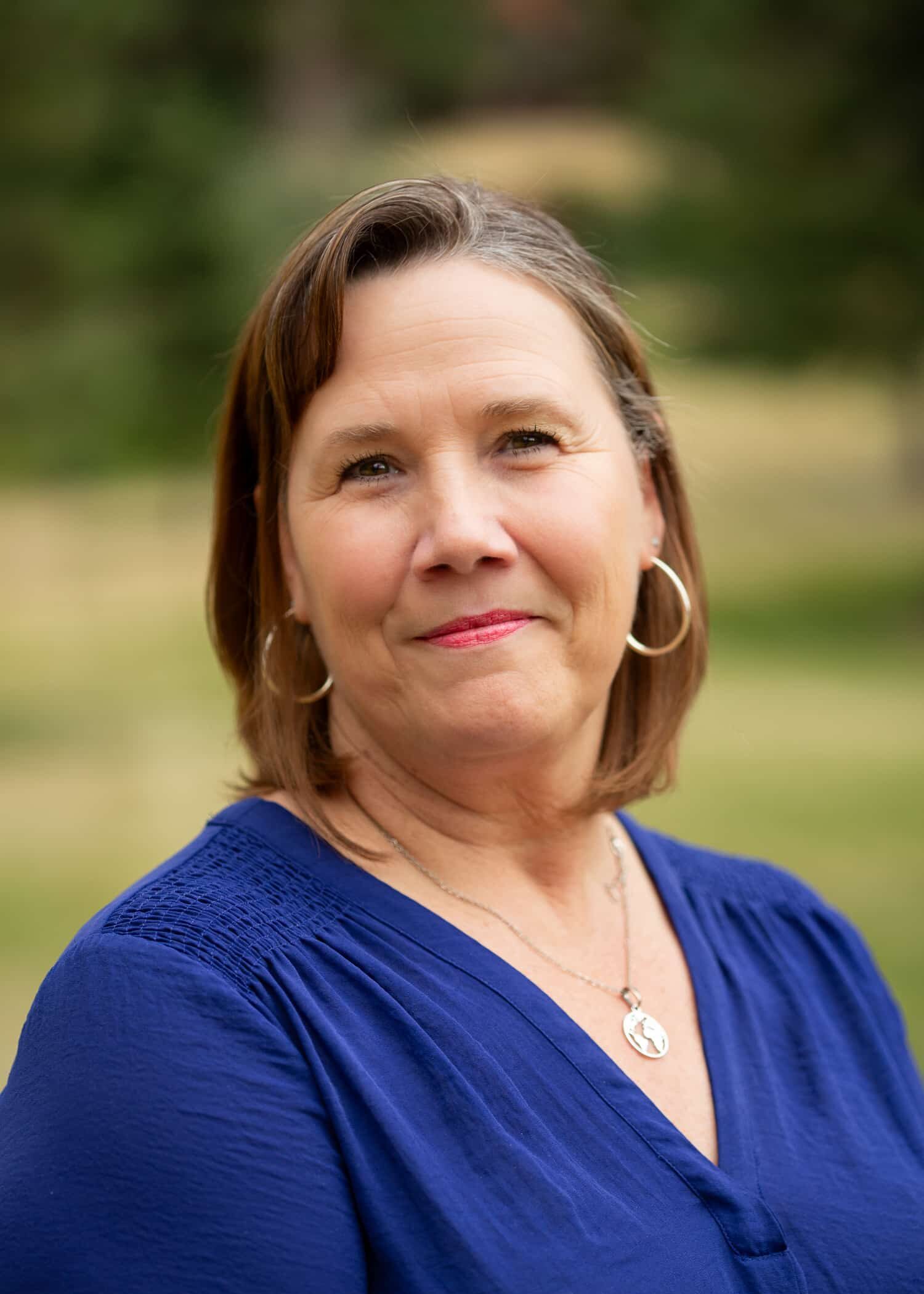 Portrait of a smiling middle-aged woman wearing a blue top, outdoors with a blurred natural background.