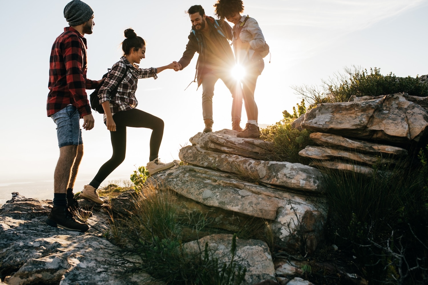A group of friends helps each other climb a rocky trail as the sun shines behind them at a mountaintop.