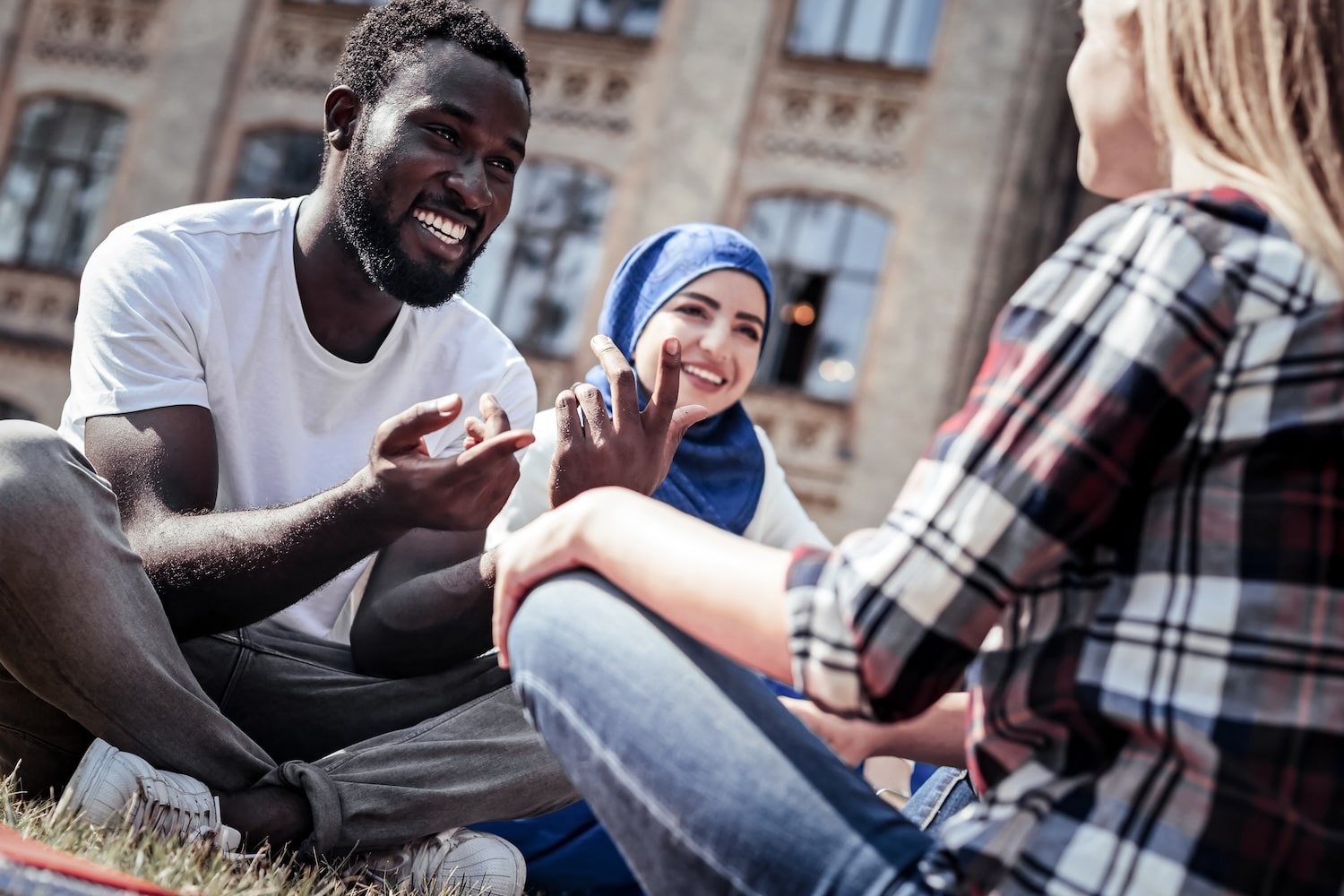 Three friends sit on the grass, smiling and chatting outdoors with a brick building behind them.