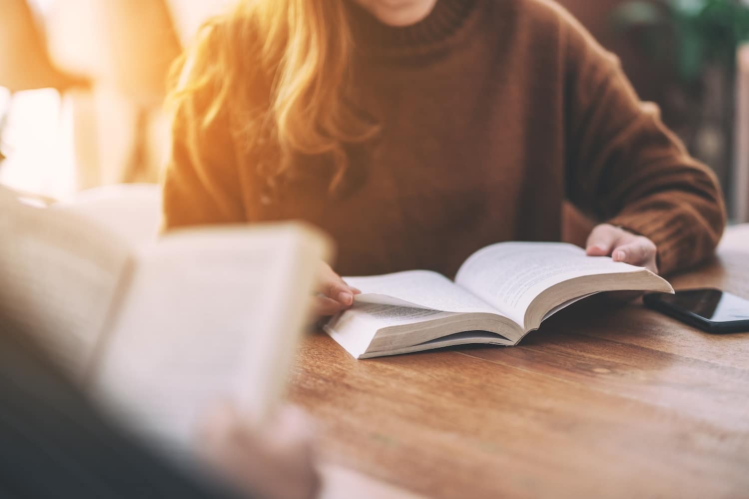 Person in a brown sweater reads an open book on a wooden table, with a smartphone nearby.