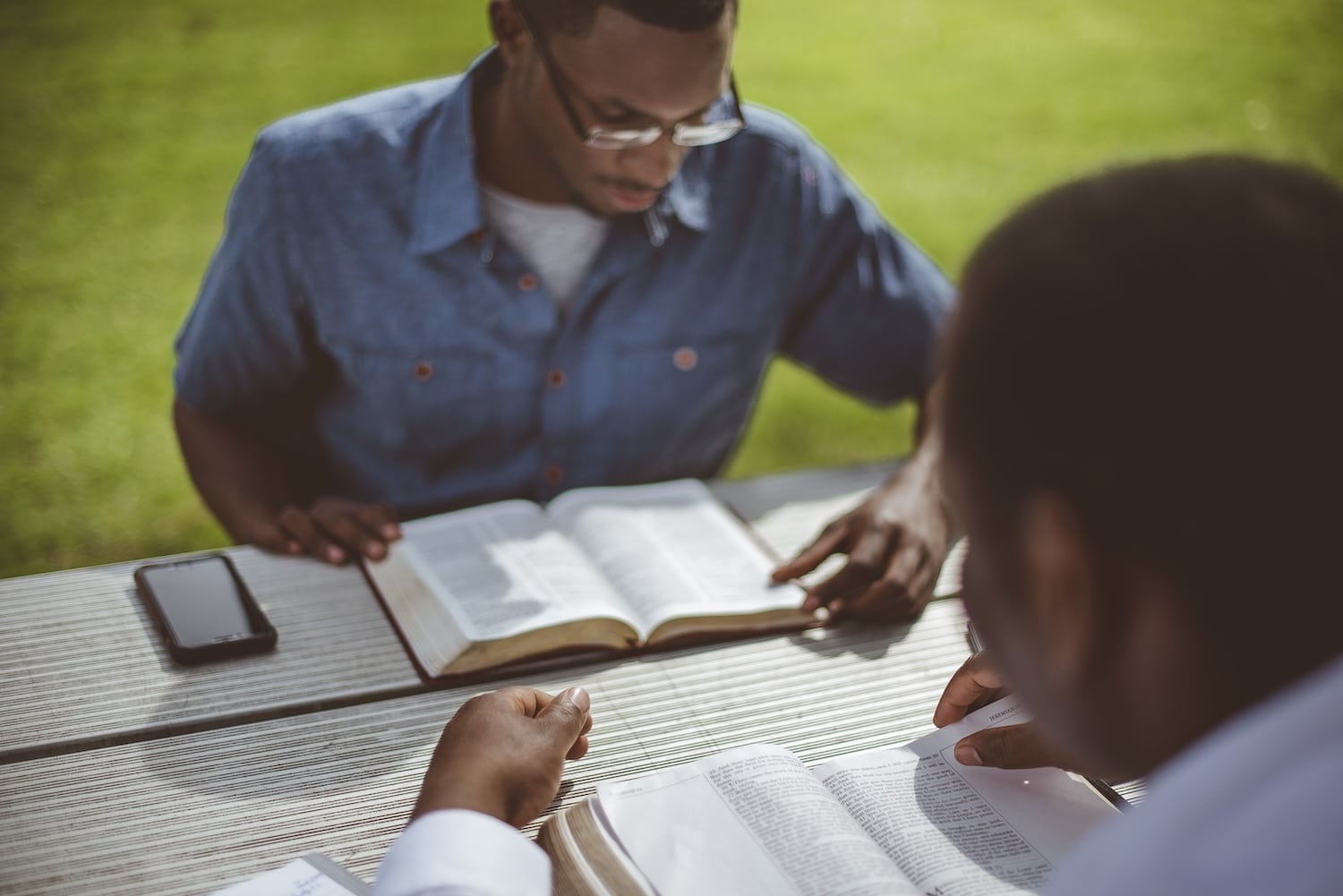 Two men study Bibles at a wooden picnic table outdoors, one in the foreground with a white shirt and the other wearing glasses and a denim shirt.