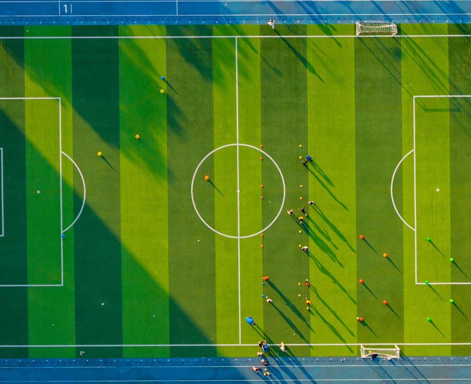 Aerial view of a soccer field with alternating green stripes, white lines, and players standing near the center circle casting long shadows.