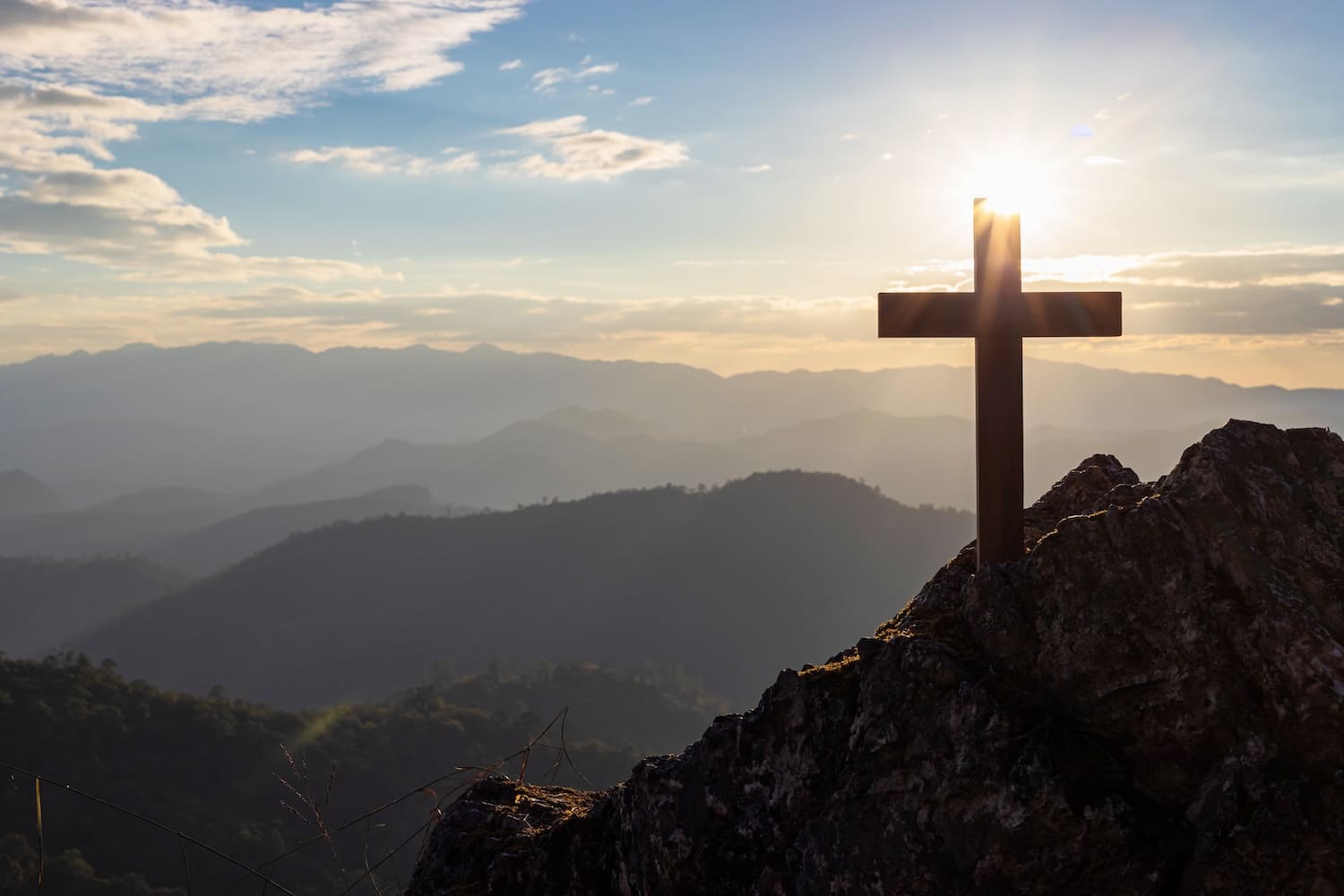 Wooden cross on a rocky peak at sunrise with misty mountain ranges in the distance.