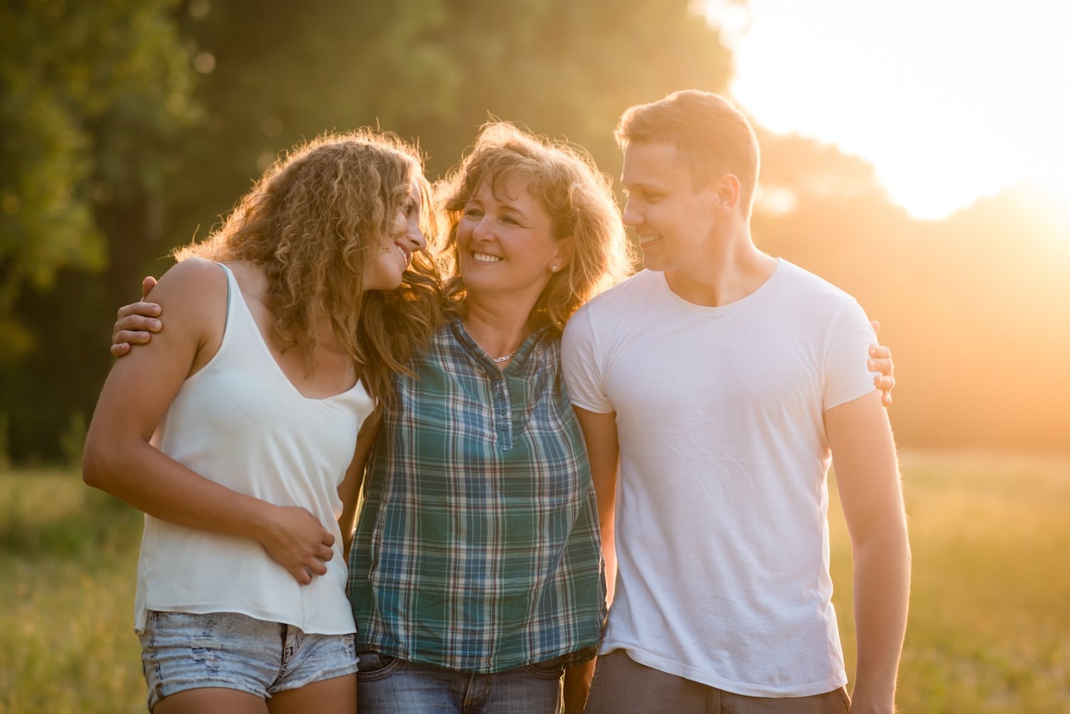 Three adults stand arm-in-arm in a sunlit park, smiling at each other during golden hour.