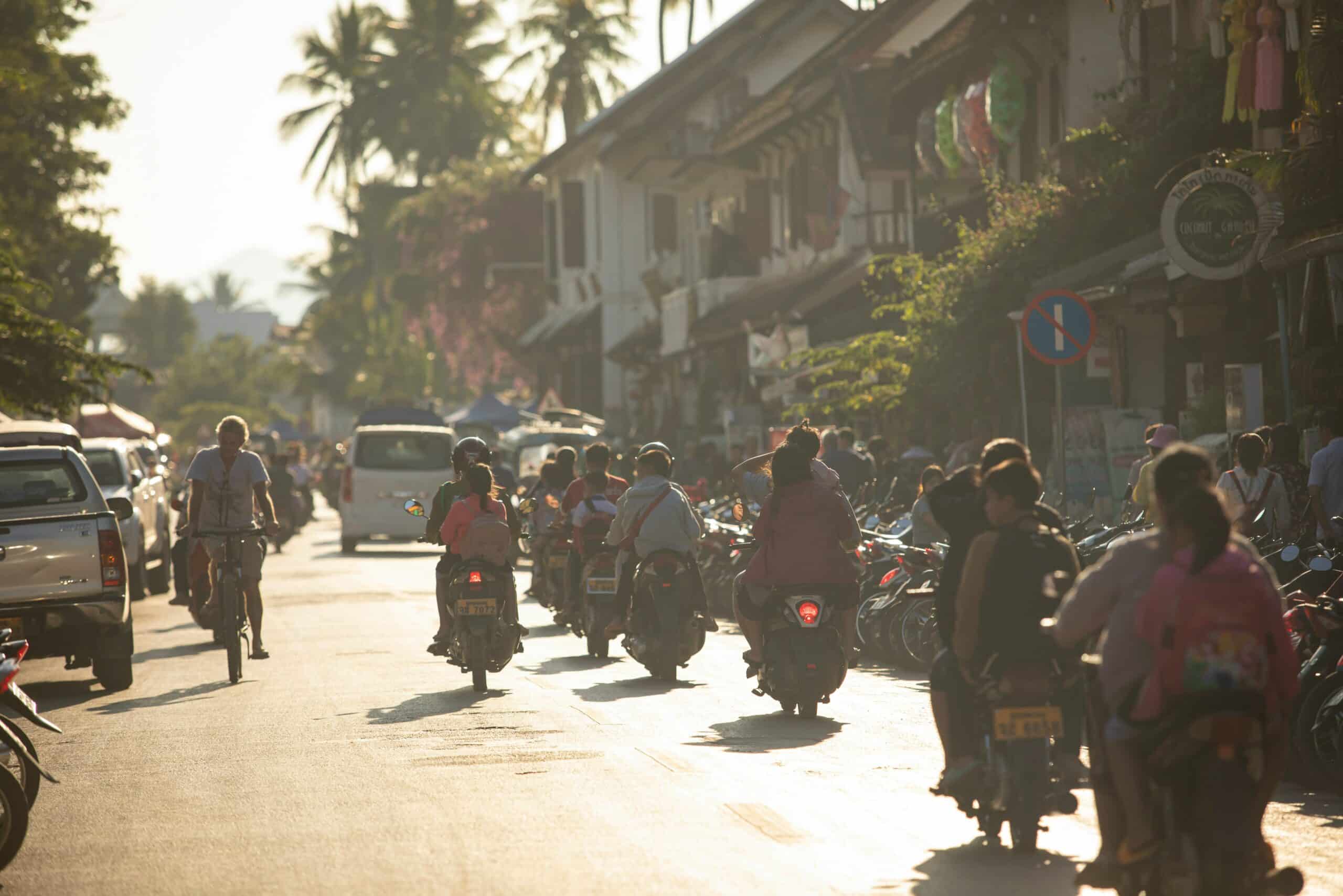 Busy street scene at golden hour with many motorbikes and pedestrians along storefronts and parked cars on the left.