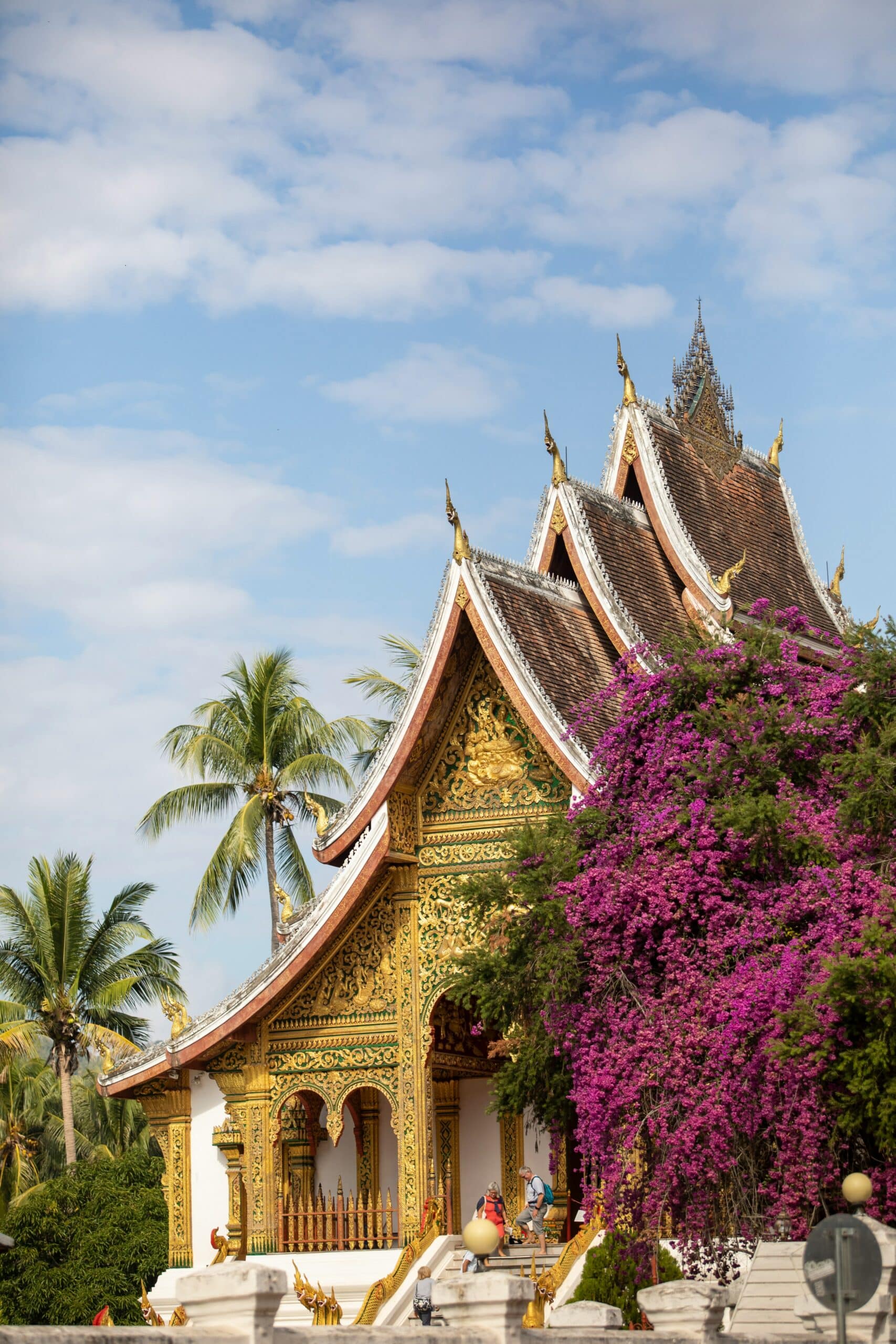 Ornate temple roof with gilded decorations, purple bougainvillea, and palm trees against a blue sky; visitors walk along the steps below.