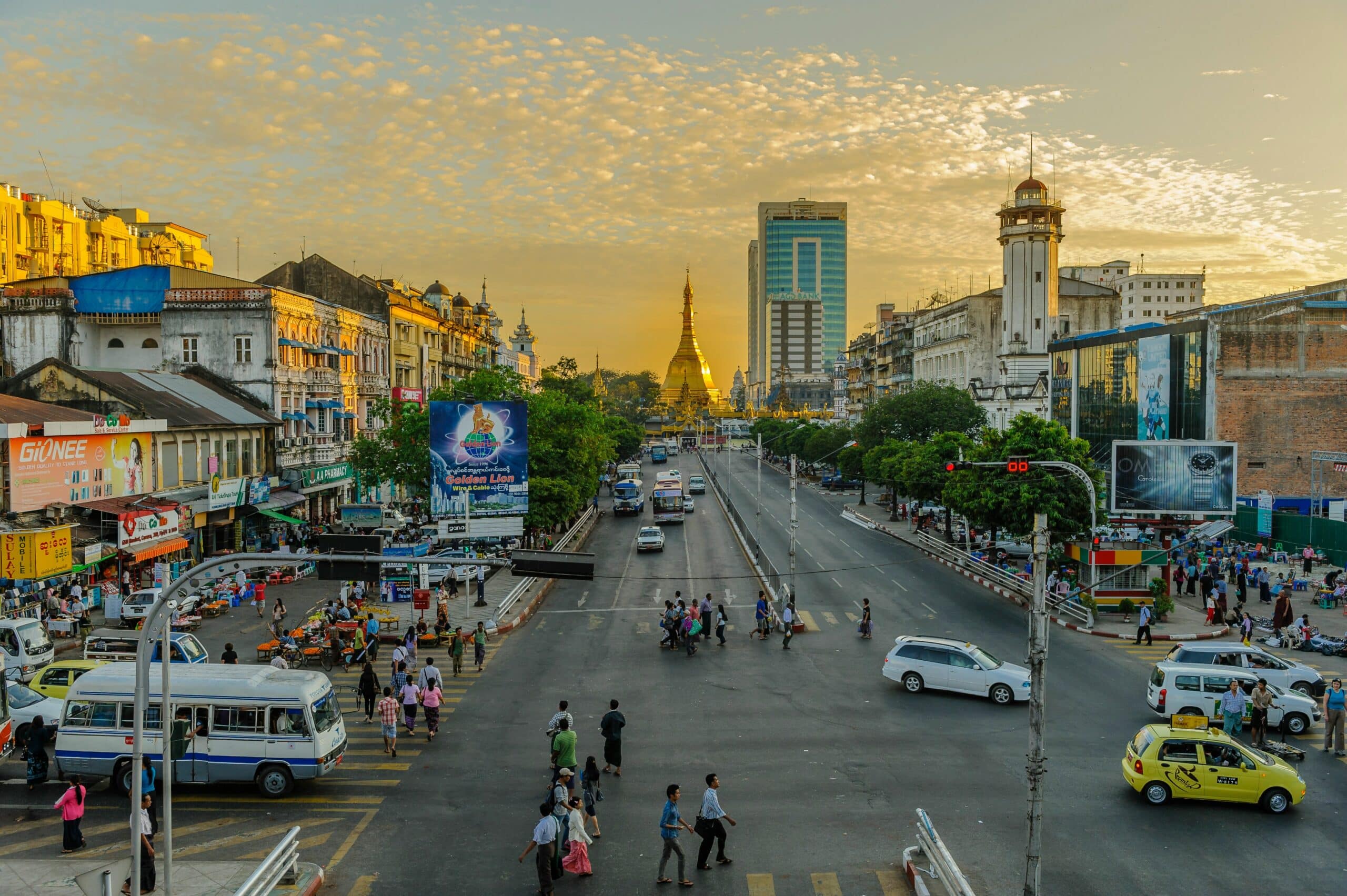 Busy city street at sunset with yellow buildings, pedestrians crossing and a distant golden pagoda rising above the skyline.