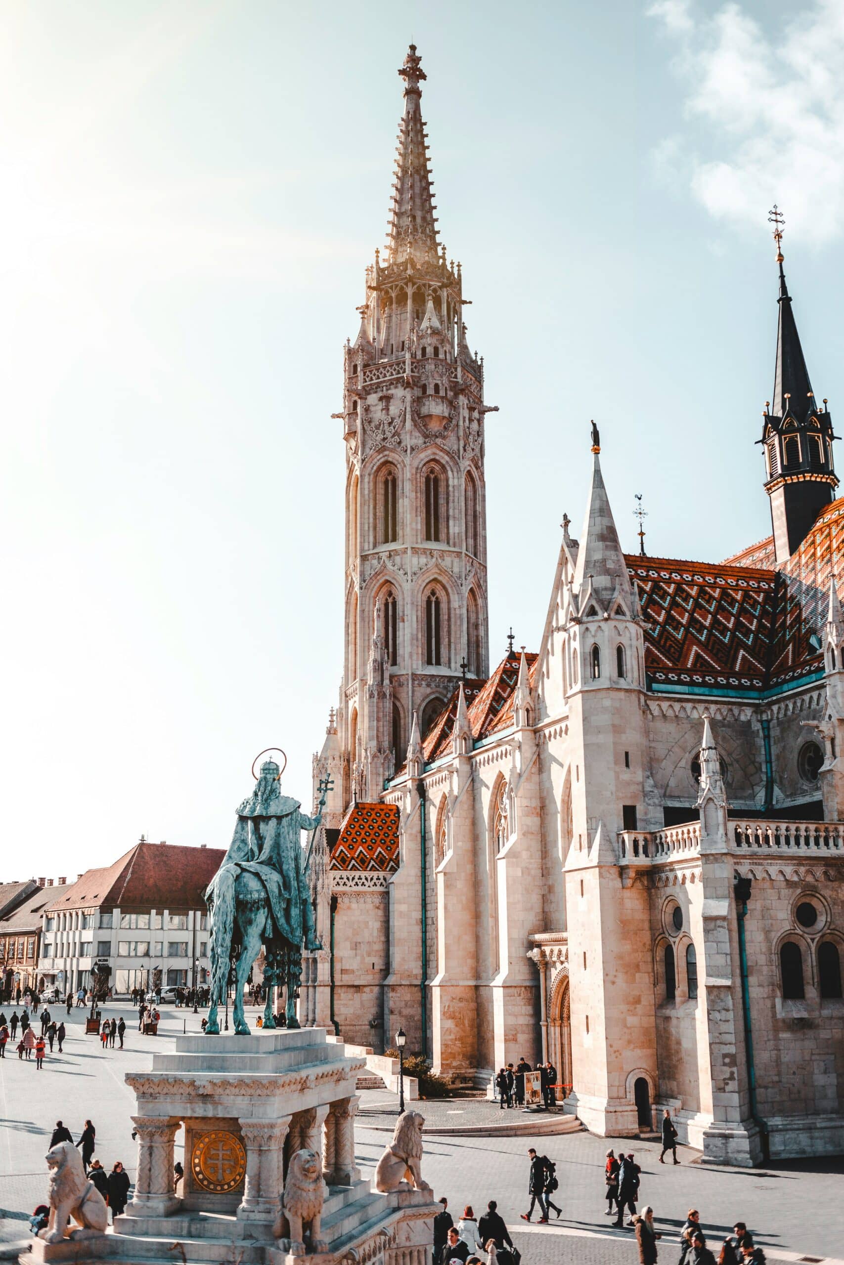 Gothic cathedral with a tall ornate spire in a sunny square, people strolling nearby.