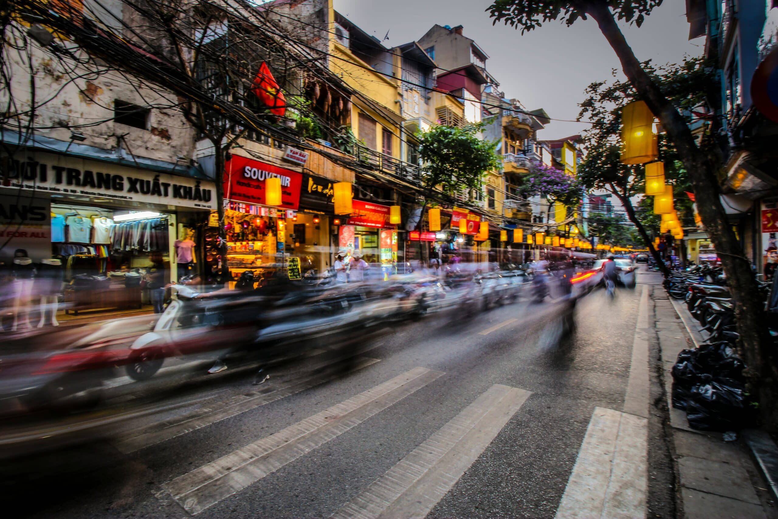 Busy urban street in Vietnam with storefronts, yellow lanterns, and blurred motorcycles rushing by.