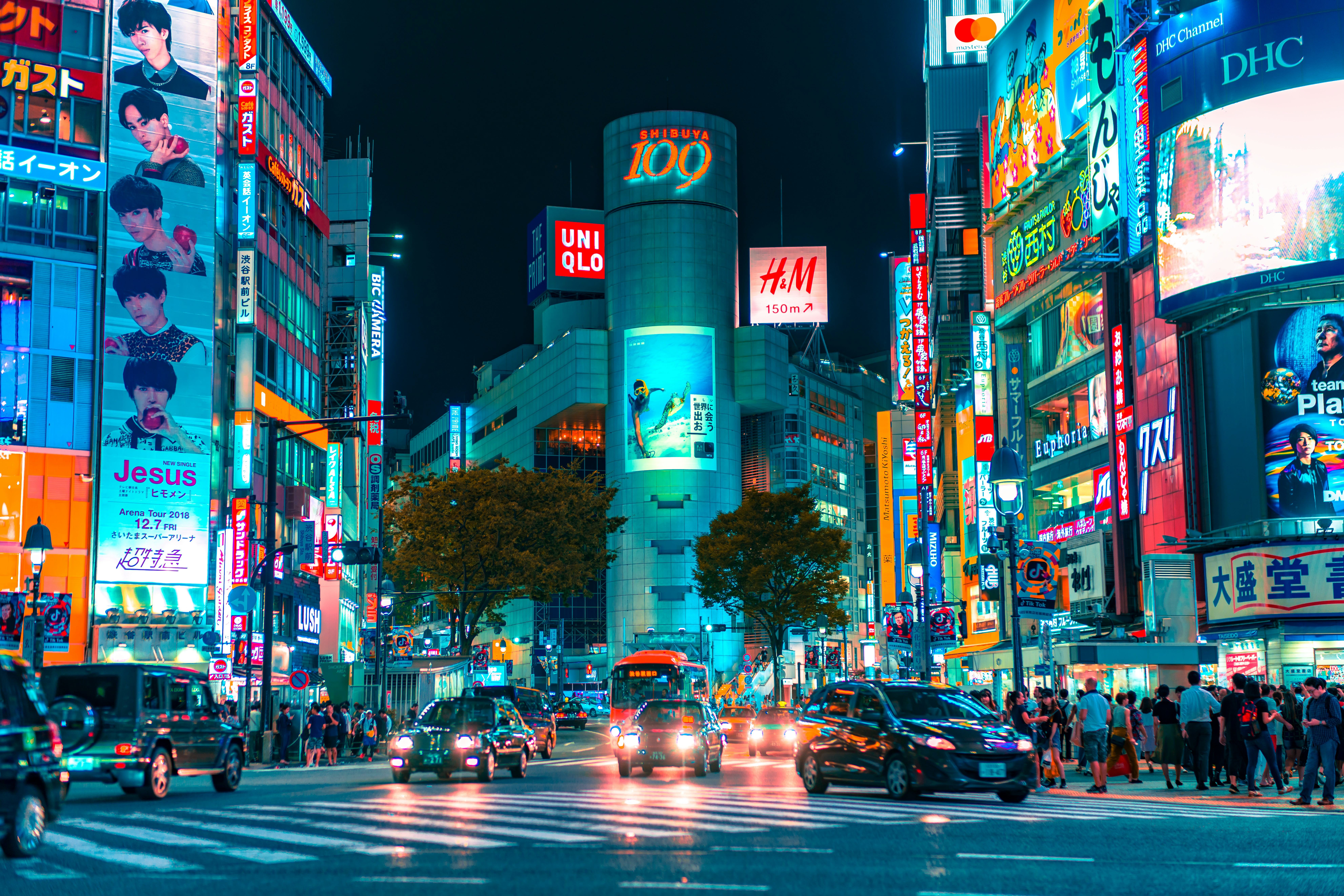 Busy Shibuya intersection at night with neon signs, crowds, and cars on a wide crosswalk area in Tokyo.