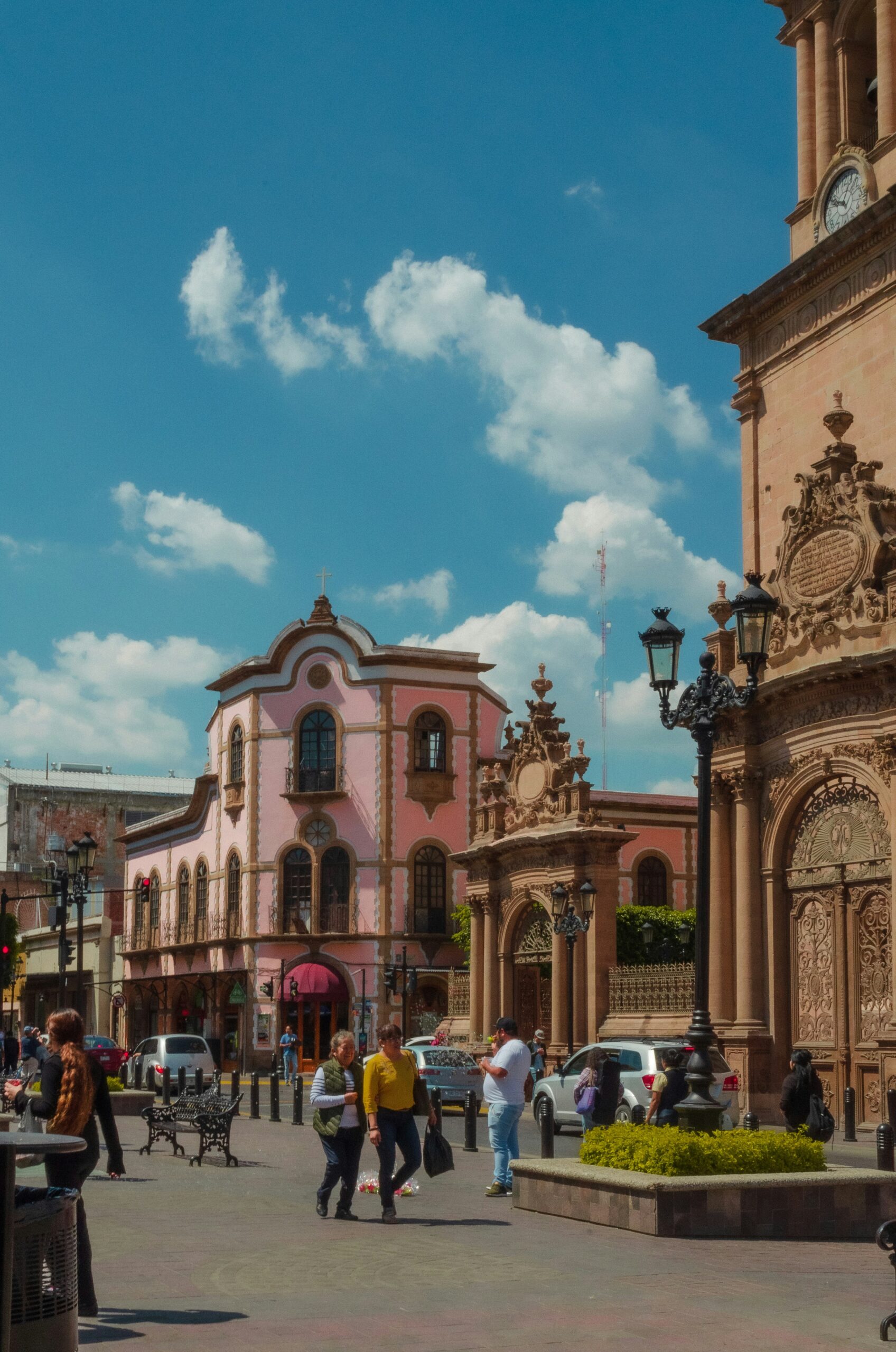Pedestrians walk in a sunny plaza beside a pink historic building with arched windows and decorative details under a blue sky.