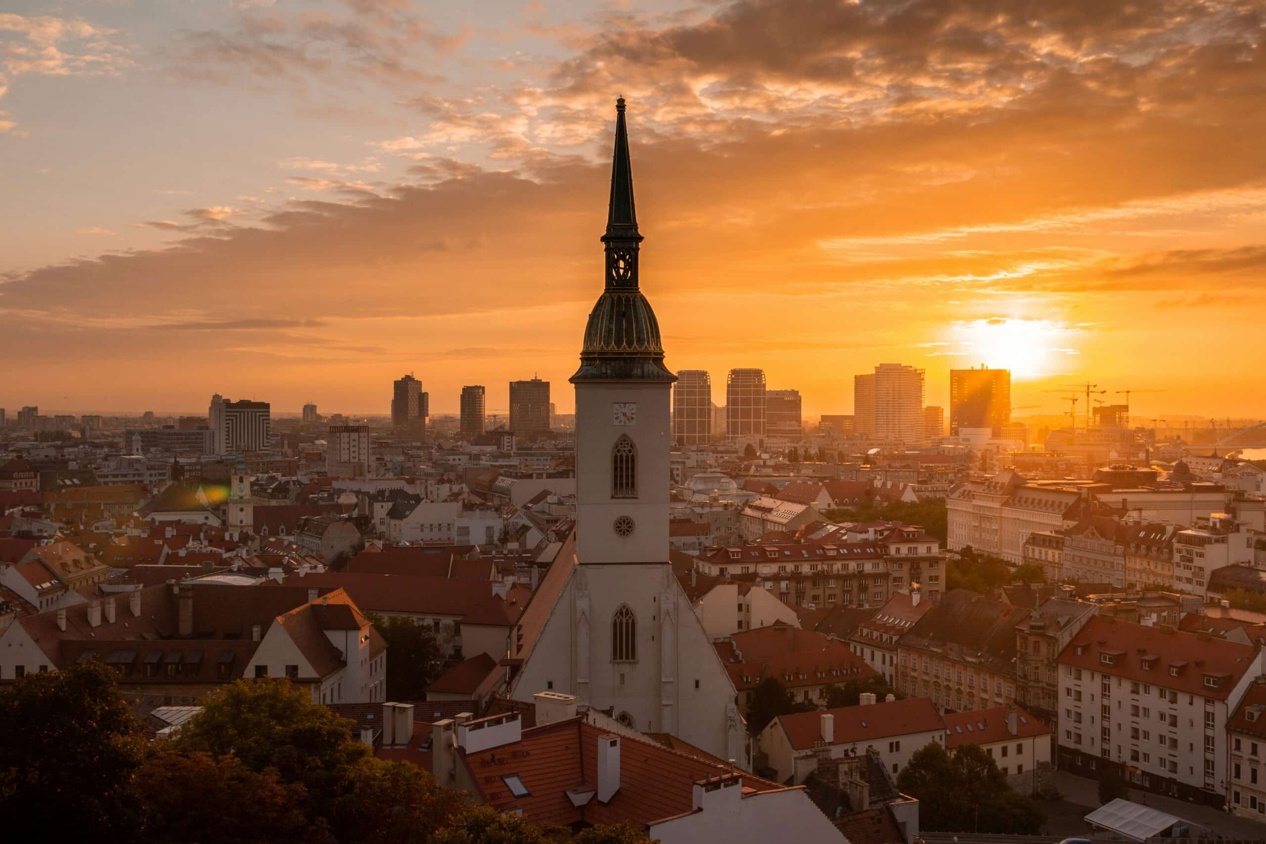 Sunset over a European city with a tall church steeple in the foreground.