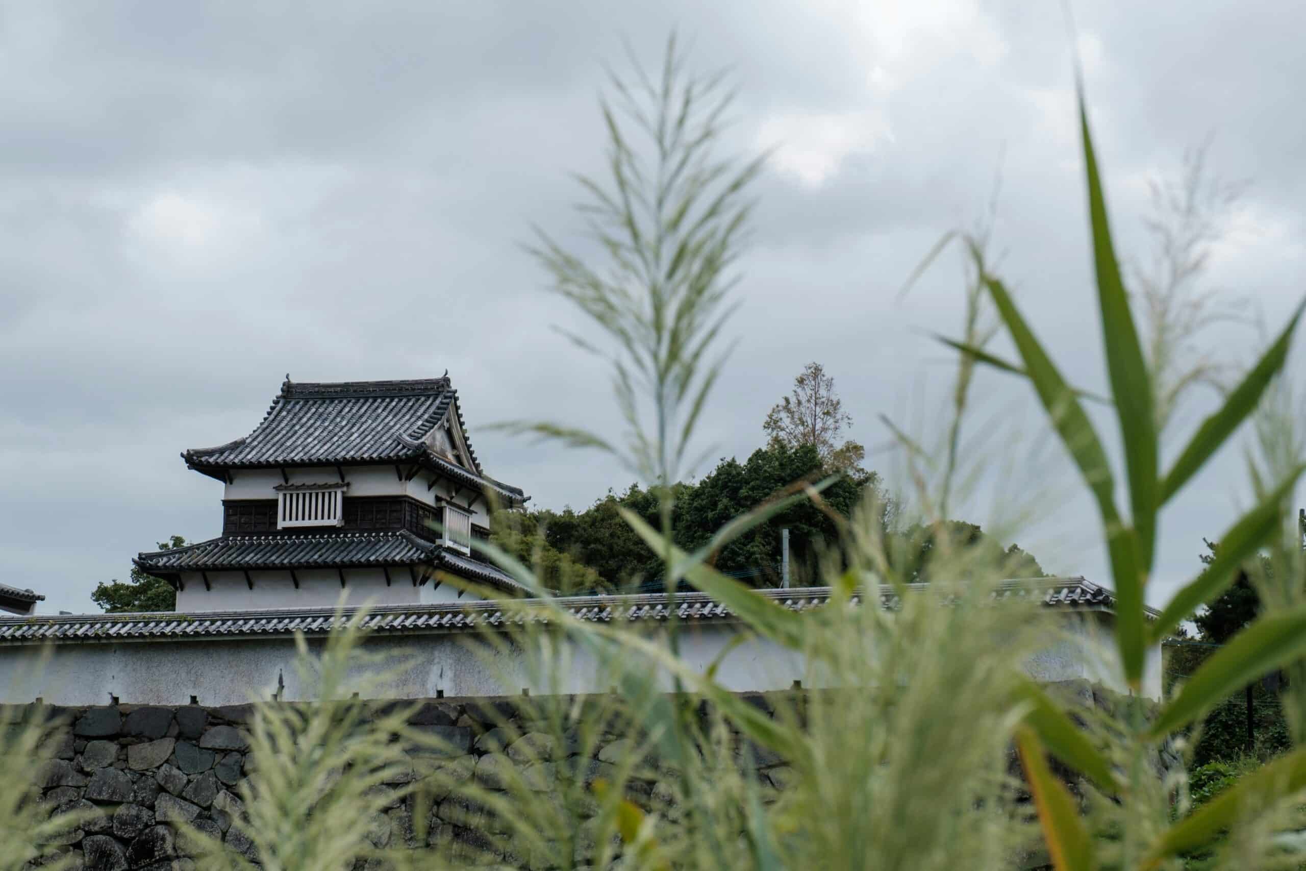 Traditional two-story tiled-roof building with white walls nestled behind a stone wall, framed by tall grasses in the foreground under a cloudy sky