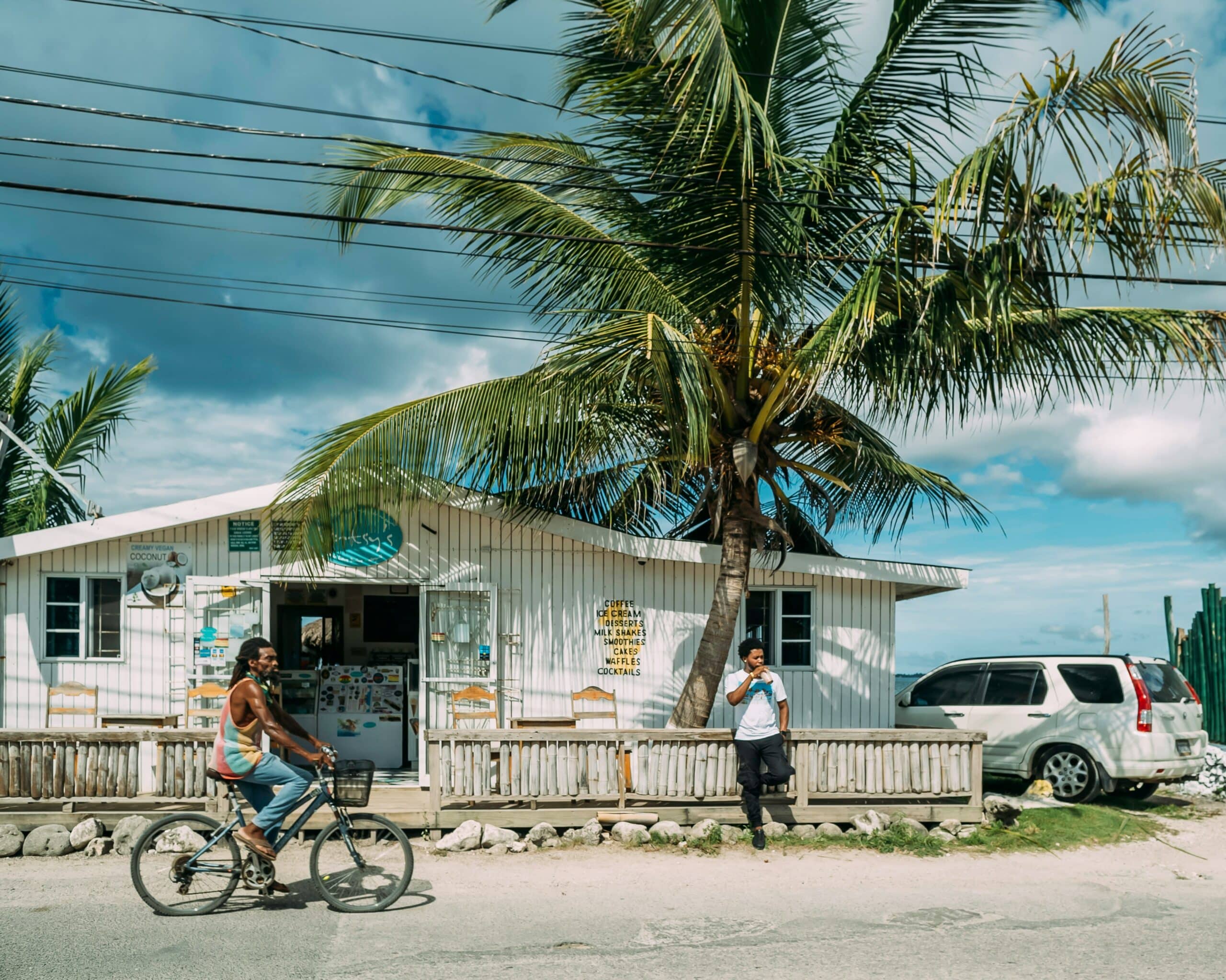 Beachfront shop with palm trees, a cyclist passing by, and a man talking on his phone beside a white building.