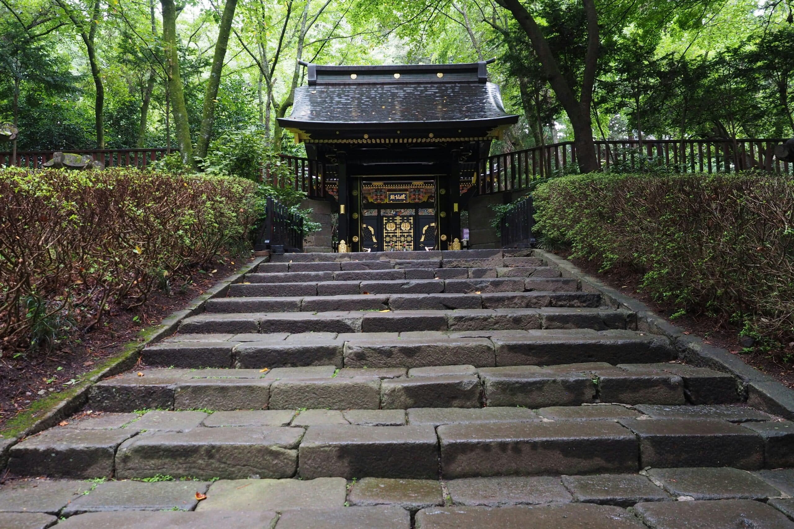 Stone steps lead up to a black and gold traditional gate framed by green trees in a garden setting.