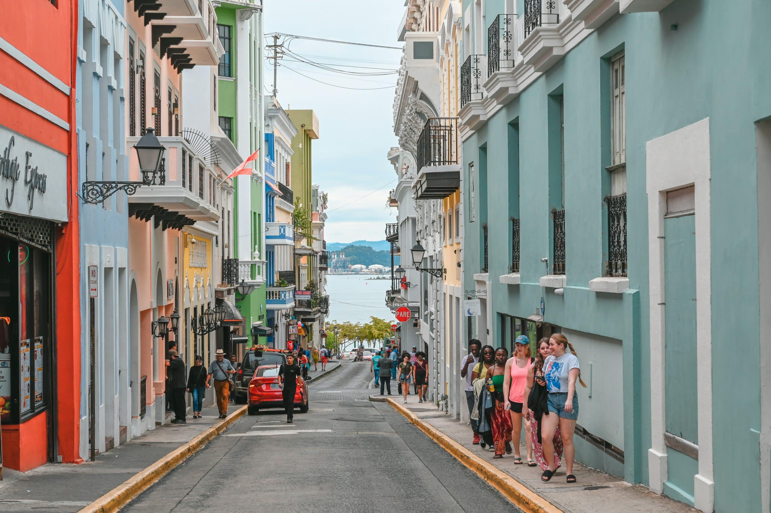 Narrow pastel-colored street lined with buildings, pedestrians walking, and a distant harbor visible at the end.